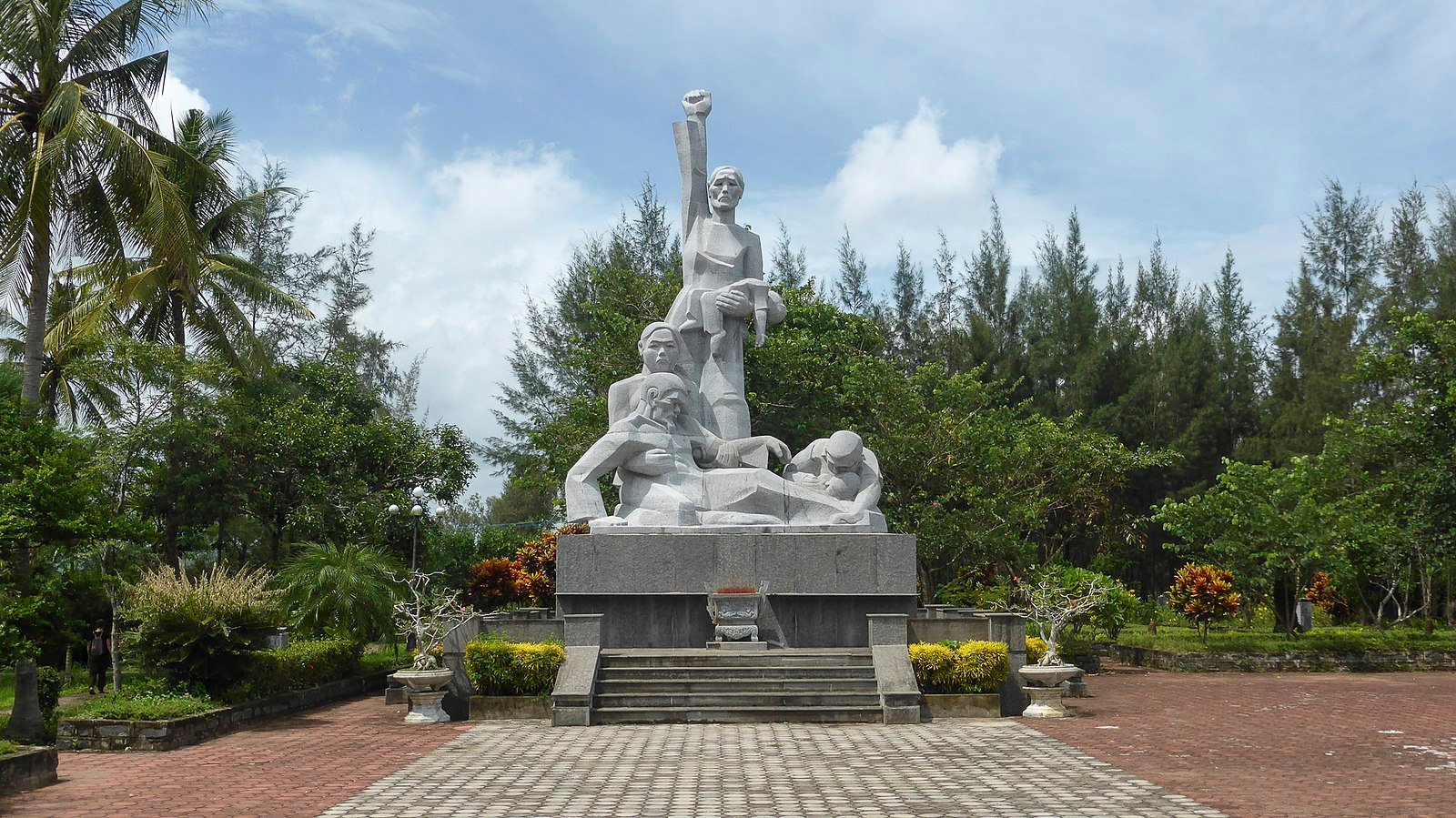 A large stone monument depicting four figures in a park setting, with one figure raising a fist. Surrounded by trees and paved path under a cloudy sky.