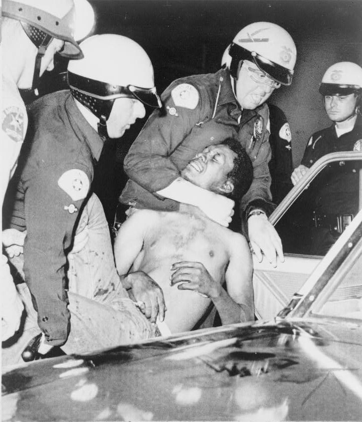 Police officers detain a shirtless man, holding him firmly, near a car. The scene appears tense, with multiple officers present.