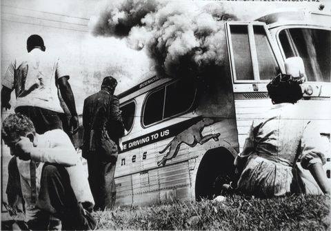 Bus engulfed in dark smoke, people stand nearby observing, others crouch on grass; an apparent emergency scene with tension and urgency.