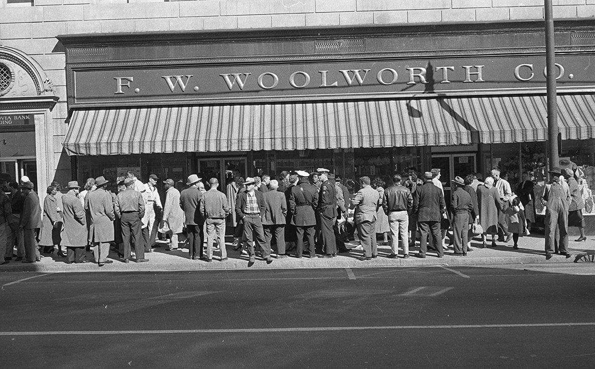 Crowd gathered outside F.W. Woolworth Co. store with striped awning; men and women in coats and hats, some speaking to uniformed officers.