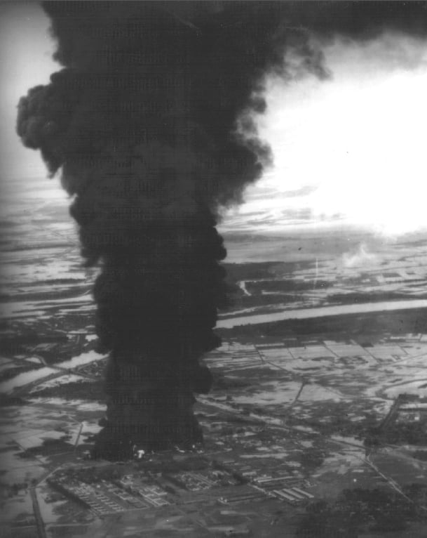Aerial view of a large, dark plume of smoke rising from a fire in an industrial area, surrounded by fields and a river in the landscape.