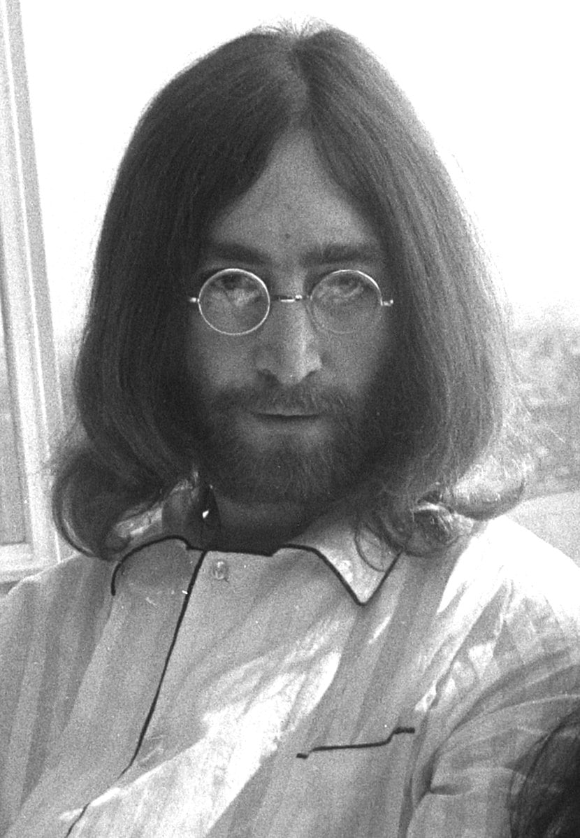 Man with long hair and round glasses, wearing a striped shirt, looking directly at the camera. The background appears to be bright and softly lit.