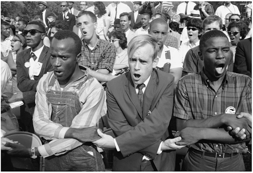 A diverse group of people, including men and women, stand closely together, holding hands and singing passionately during a civil rights protest.