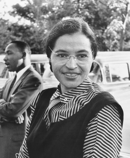Woman in glasses with striped blouse smiling, outdoors. A man stands behind, near vintage car. Trees in the background. Black and white photo.