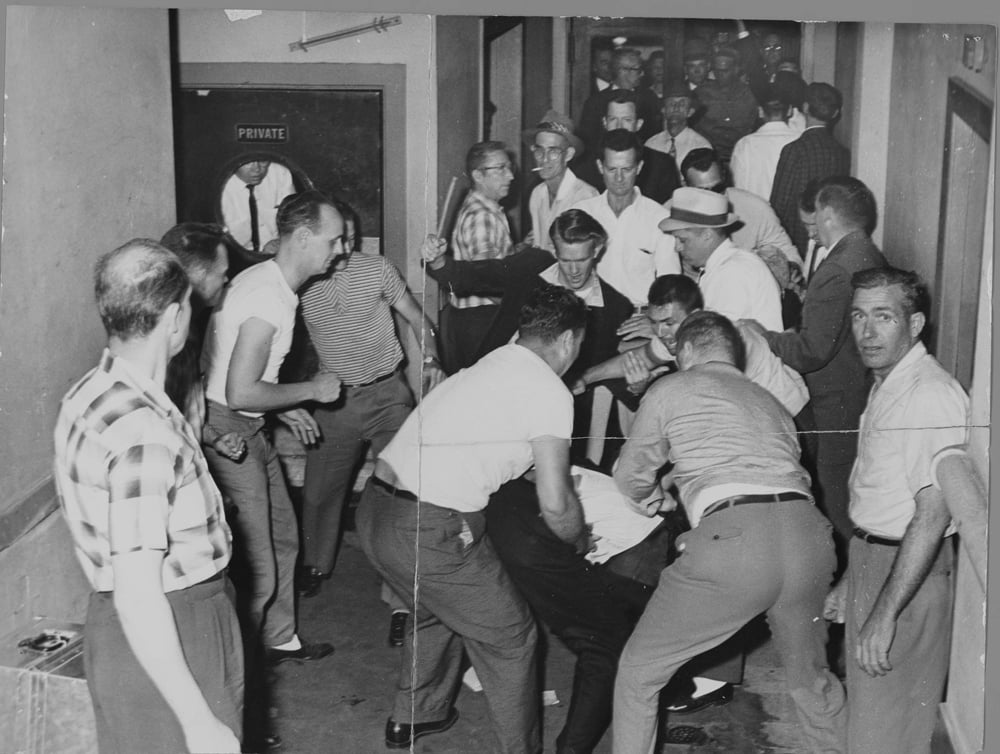 Crowd of men in a narrow corridor, some appear to be in a scuffle. One man lies on the floor while others gather around. A "Private" door is visible.