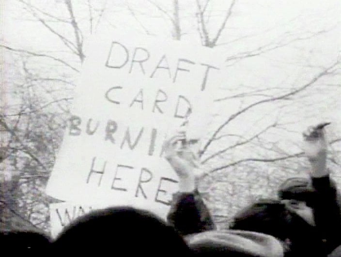 Protesters holding a sign that reads "Draft Card Burning Here" amidst a crowd, with bare trees visible in the background.