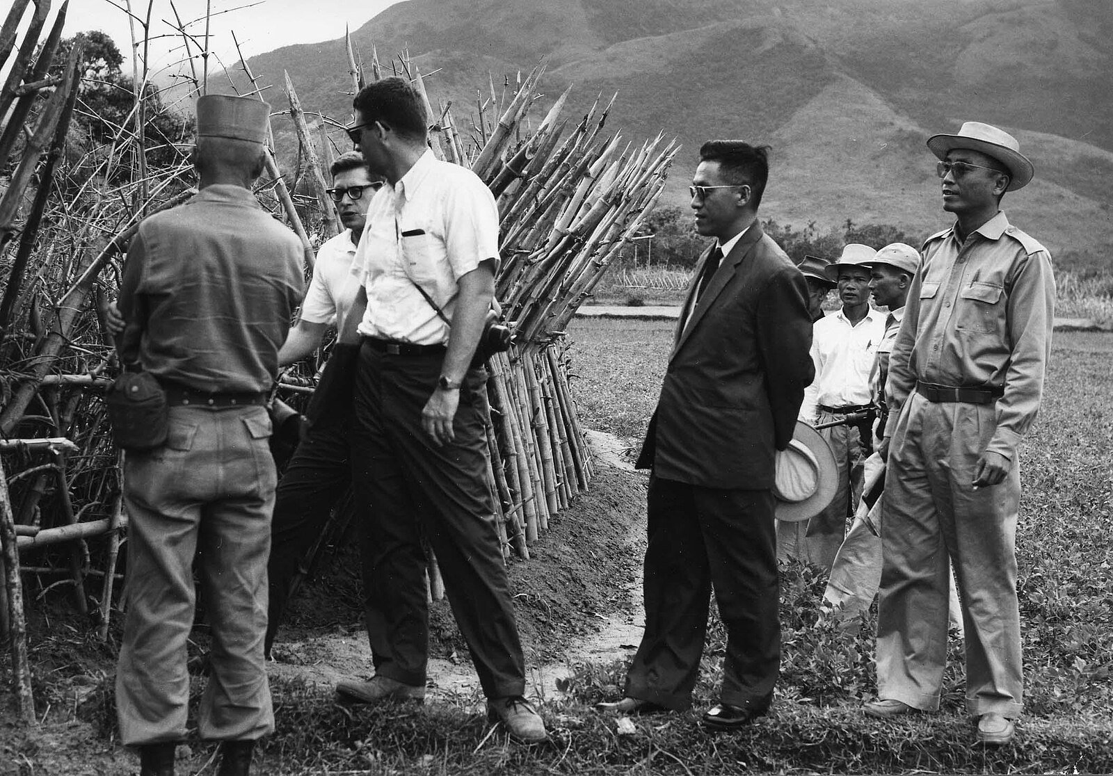 A group of men, some in military and others in civilian clothes, stand near a bamboo structure in a field with mountains in the background.