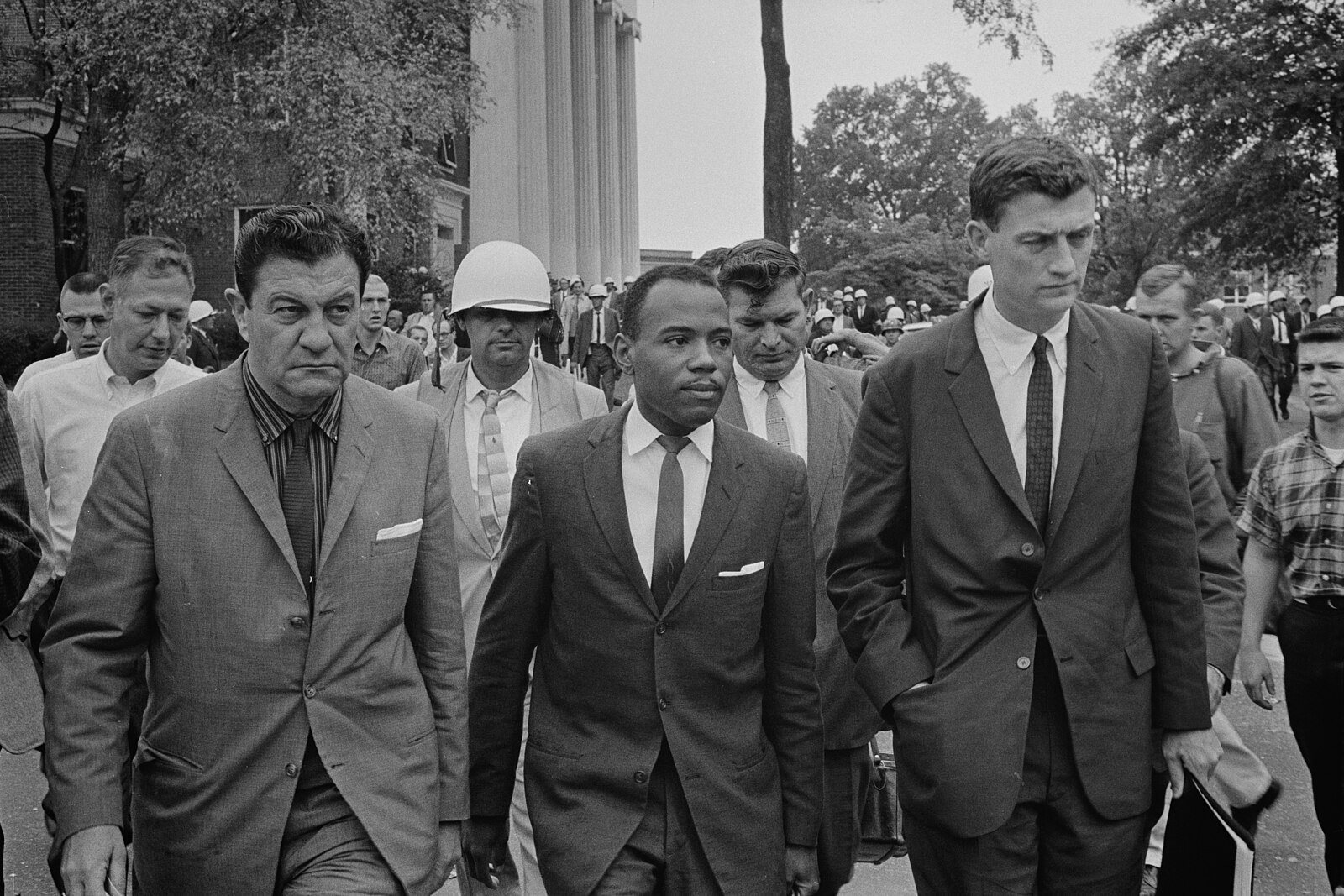 A group of men in suits walk together, with one man in a helmet. They are outside, and there are trees and a building with columns in the background.