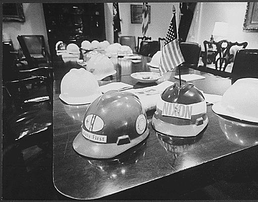 Hard hats on a long table in a conference room, one labelled "Nixon" with an American flag and another with "Safety First", chairs in the background.