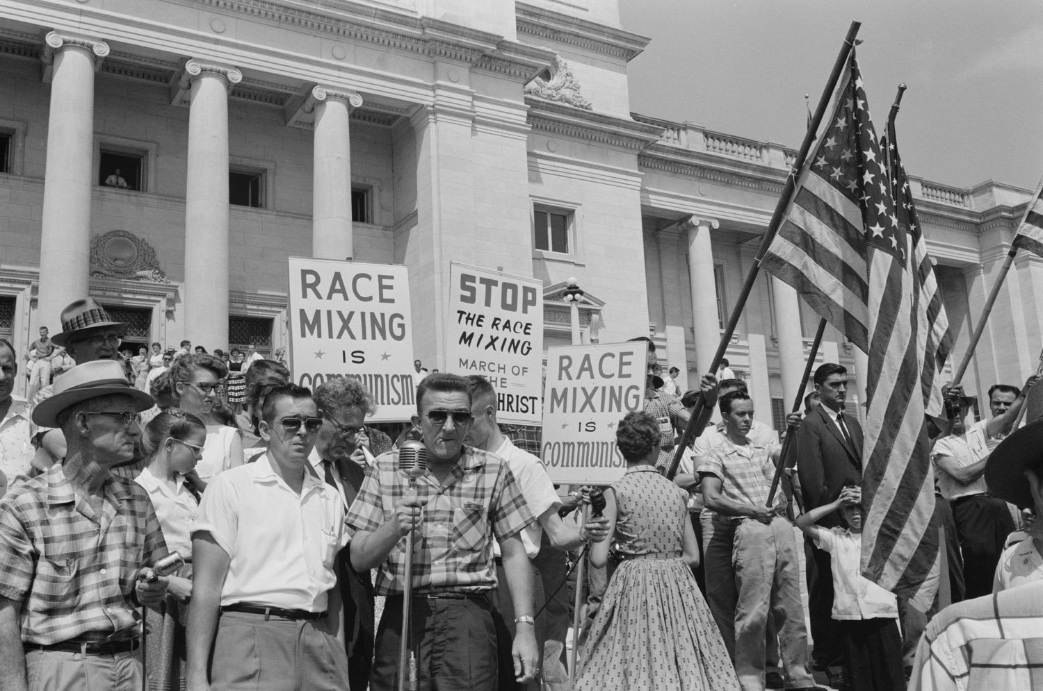 A 1950s protest outside a neoclassical building with people holding signs against racial integration, some carrying American flags.