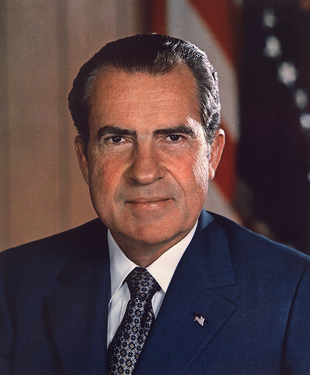 A man in a dark suit and patterned tie is seated, with an American flag visible in the background, looking forward with a neutral expression.