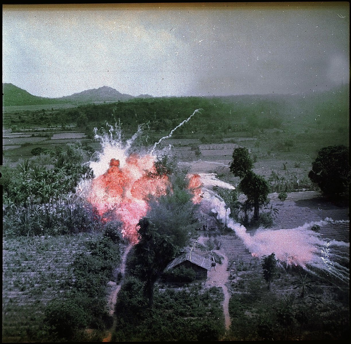 Aerial view of a rural landscape with an explosion near a hut surrounded by trees, emitting a bright flash and smoke trails. Hills in the background.
