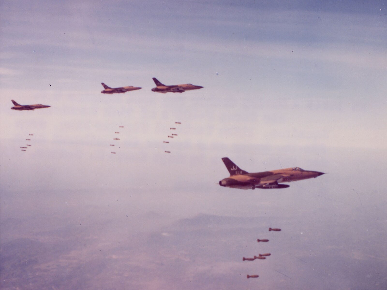 Four military jets are flying in formation over a landscape, releasing bombs that drop in a diagonal pattern towards the ground below.