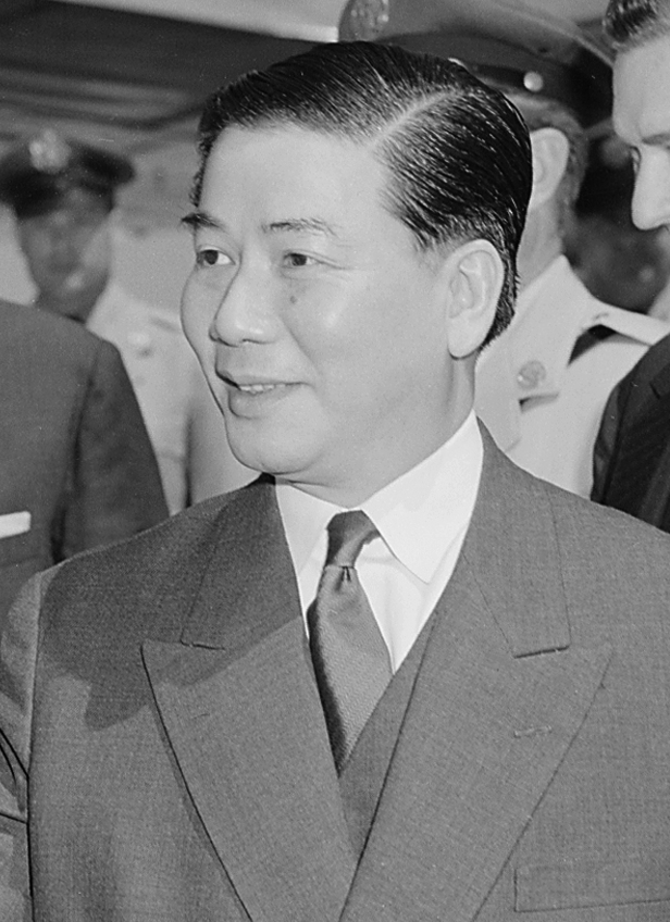 A man in a suit smiles, surrounded by others in uniform. The image is in black and white, suggesting a formal or historical context.