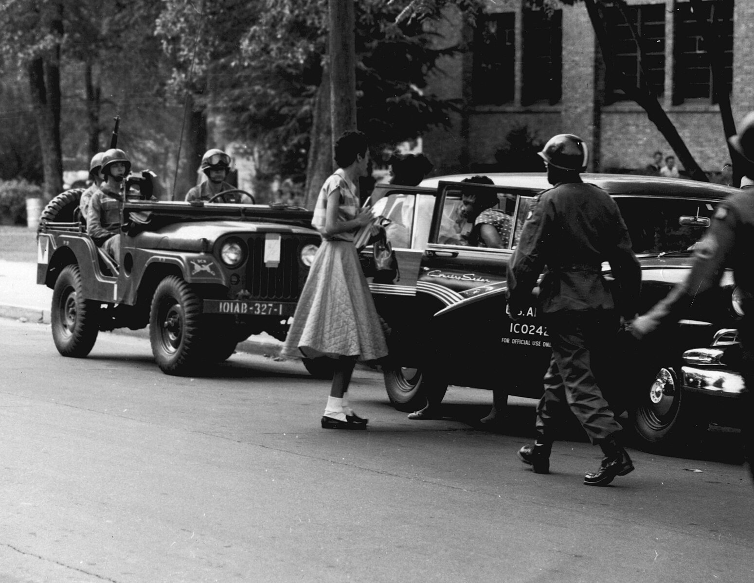 Soldiers escorting a young woman past a military jeep and car in a street setting, with trees and a brick building in the background.