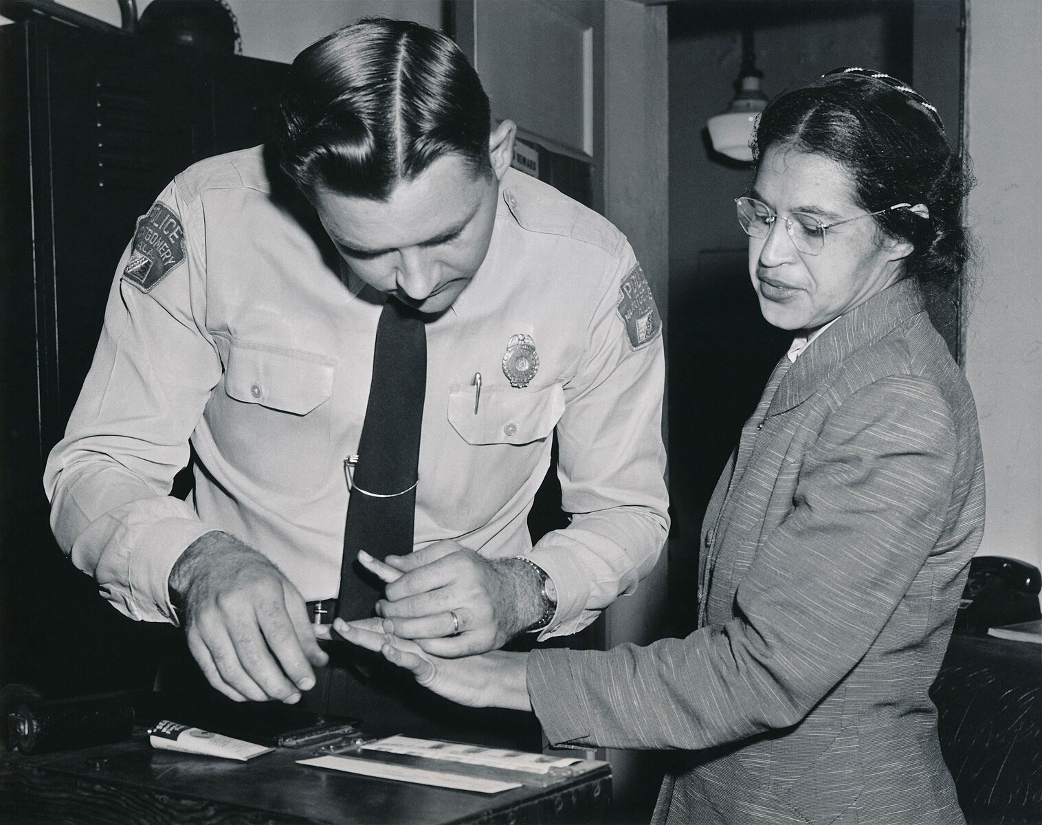 A policeman takes fingerprints of a woman in a formal suit in an office setting. She stands calmly while he focuses on the task.