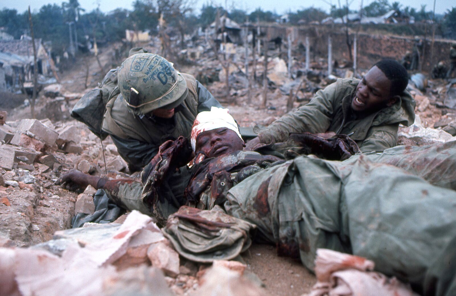 Soldiers assist an injured comrade lying amidst rubble. The scene is chaotic with debris and damaged buildings in the background, indicating a recent battle.