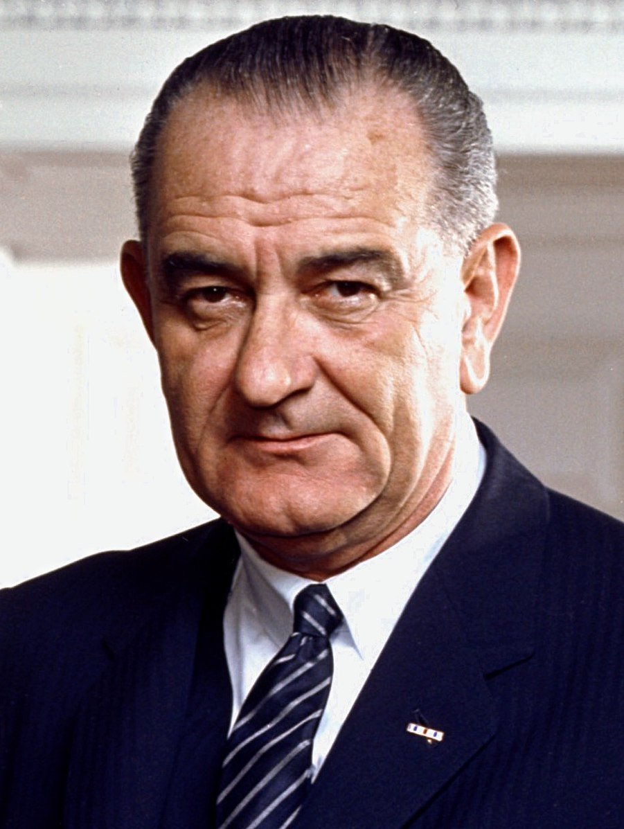 Elderly man in a dark suit with a striped tie, looking directly at the camera with a neutral expression, background is a light-coloured wall.