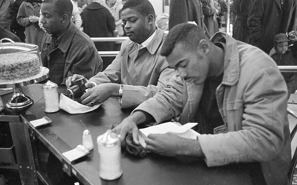 Three men sit at a lunch counter with cameras in front of them, appearing focused and calm. A cake is displayed on the counter nearby.