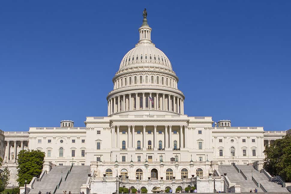 United States Capitol building with a prominent dome and classical columns under a clear blue sky, surrounded by trees and steps leading up to the entrance.