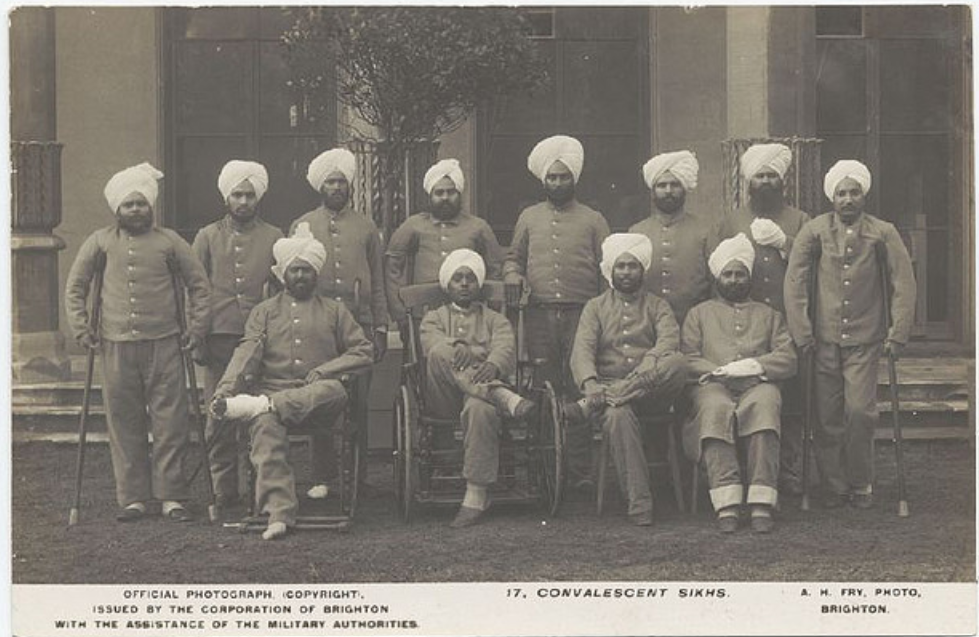 Sepia-toned photograph of a group of Sikh soldiers in convalescence during the First World War. The men are dressed in military hospital uniforms and wearing turbans. Some are seated, including two in wheelchairs and others with their legs bandaged or in slings, while others stand behind them. Crutches are visible, indicating injuries. The group is posed in front of a building, possibly a hospital or convalescent home. The caption below reads "17. CONVALESCENT SIKHS," and credits the photograph to A. H. Fry, Brighton, issued with the assistance of the military authorities.