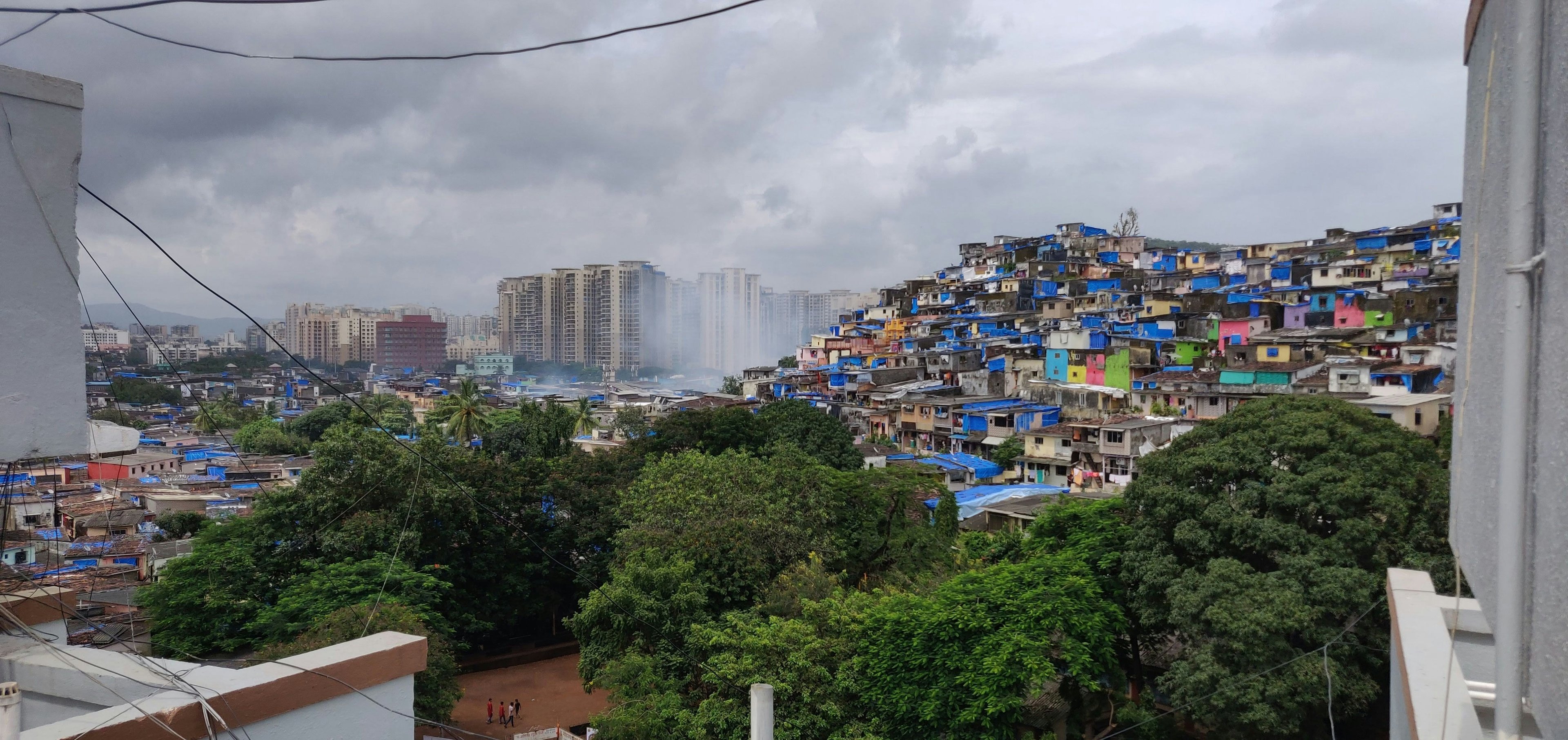 The sprawl of the Dharavi slum extends along a hillside, which is overshadowed by tall modern buildings in the background.