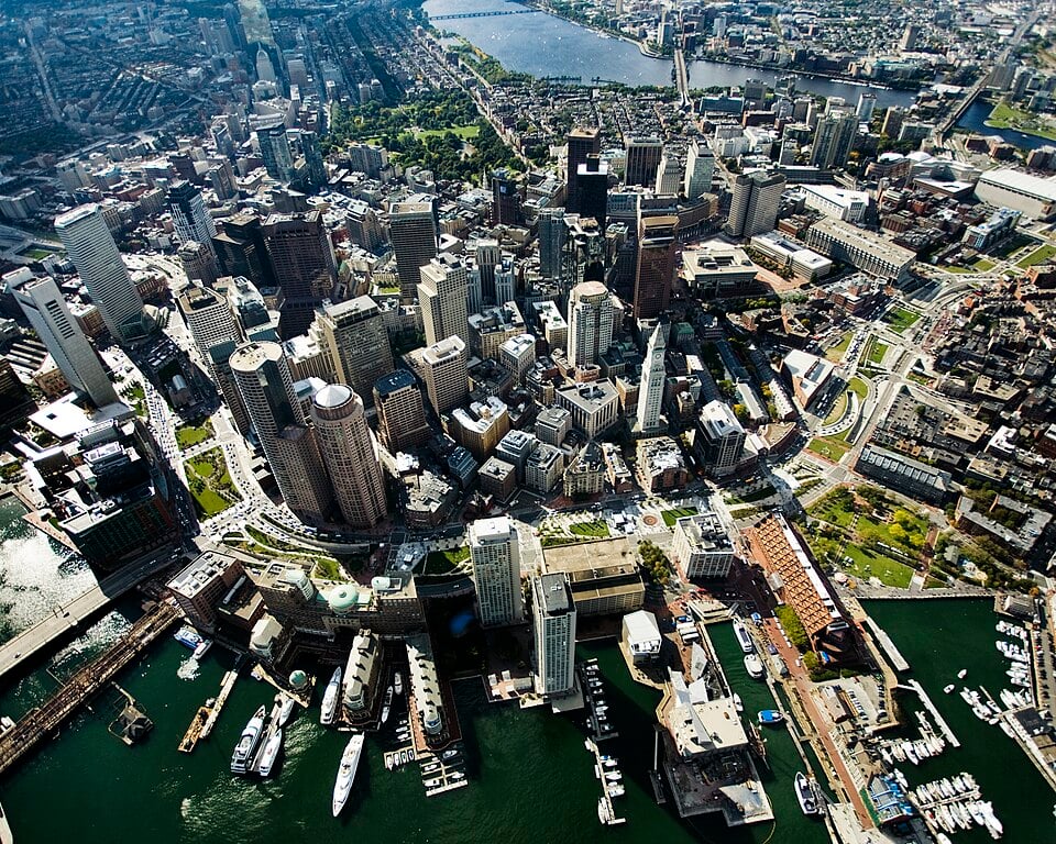 Aerial view of a bustling cityscape with modern skyscrapers, green spaces, and a river. Boats are docked in the harbour in the foreground.