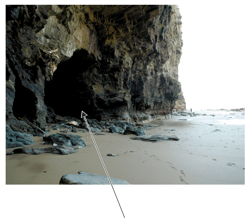 Rocky coastal cave with a pointed arrow marking the entrance. Sandy beach with scattered rocks and footprints; a person in the distance near the shore.