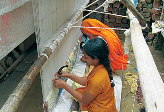 Woman in orange saree weaving a carpet on a loom outdoors, using a tool to work yarn, surrounded by loose threads and brick walls.