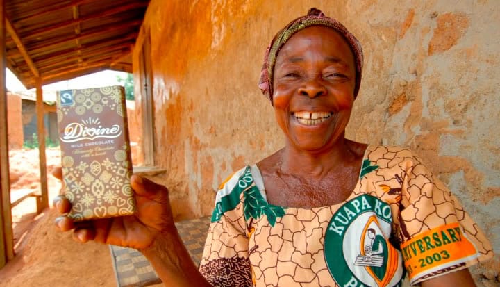 Elderly woman smiling and holding a Divine chocolate bar. She wears vibrant traditional clothing outside a building with a corrugated roof.