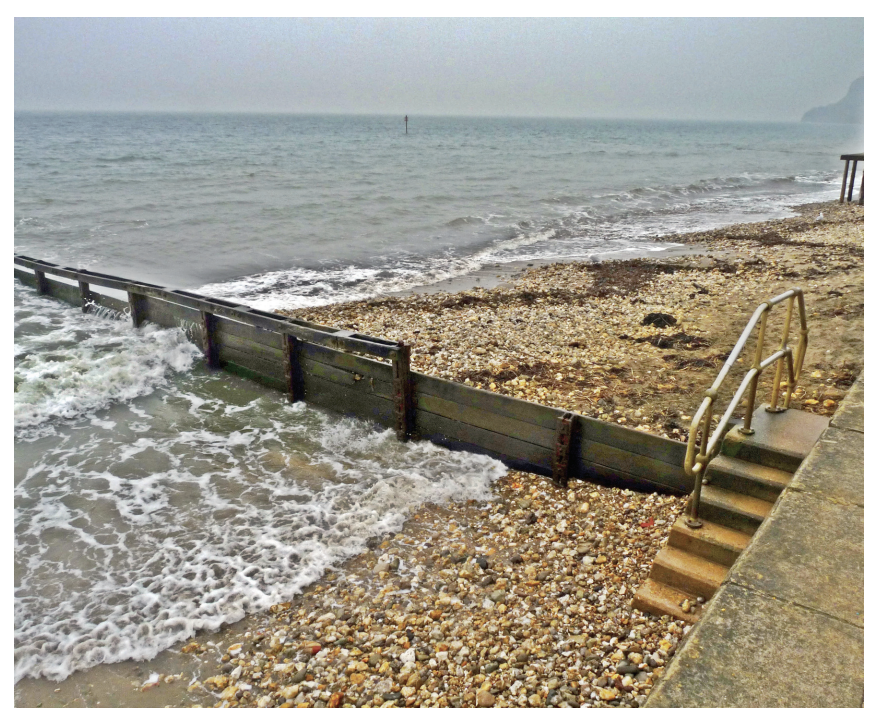 Pebble beach with a wooden groyne, concrete steps, and metal handrails leading into the sea. Overcast sky and gentle waves breaking onshore.