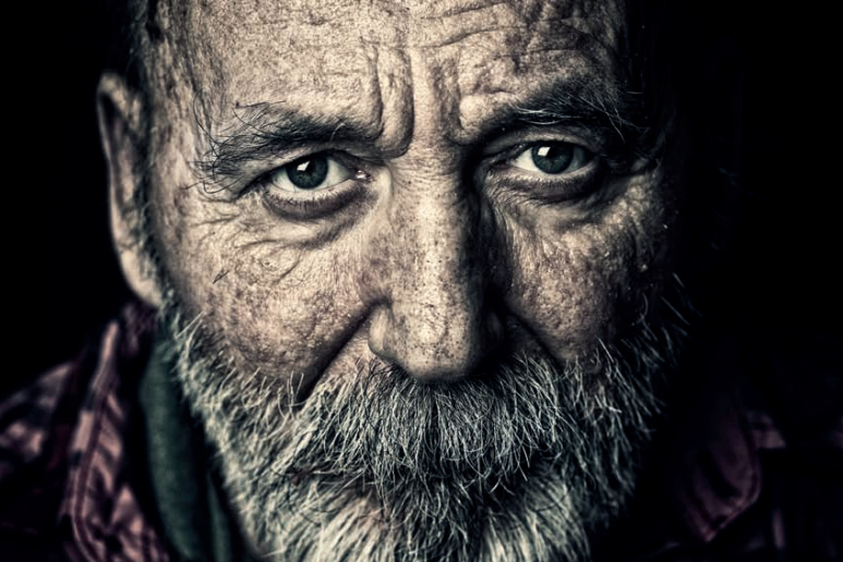 Close-up of an elderly man with a white beard and weathered skin, gazing intently. The background is dark, highlighting his facial features.