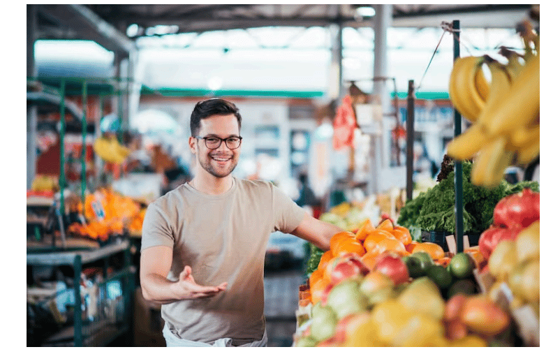 Man with glasses smiling at a vibrant market stall, surrounded by assorted fruits like bananas, oranges, and apples under a covered roof.