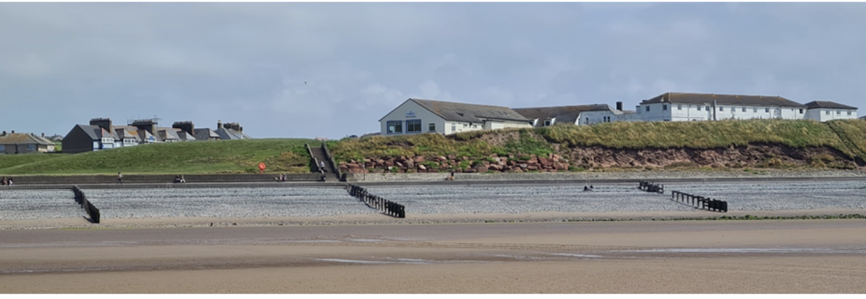 Wide sandy beach with a stone jetty leading to grass-covered cliffs topped by houses under a partly cloudy sky.
