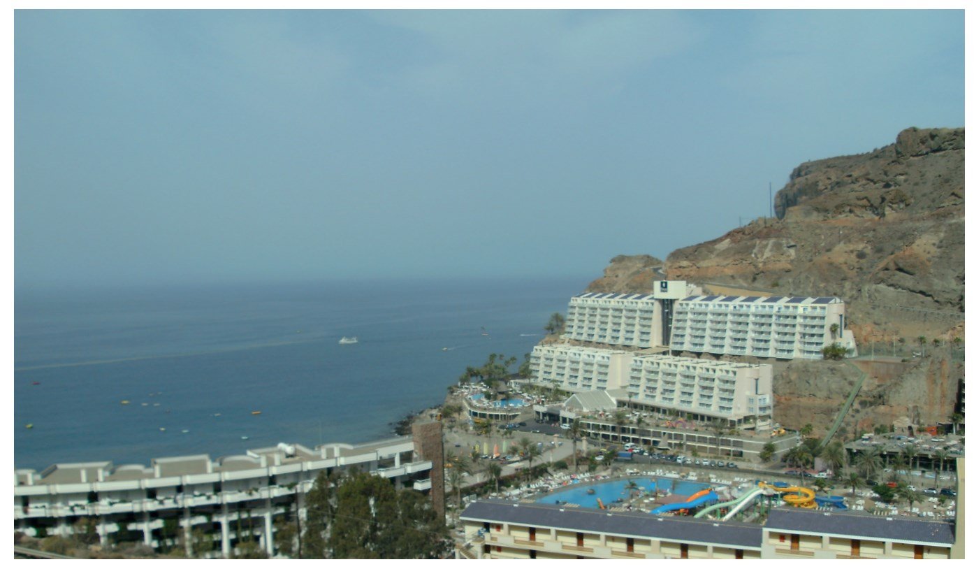 Coastal view of a resort with multiple buildings, a large swimming pool with water slides, and rocky cliffs adjacent to the ocean under a clear sky.