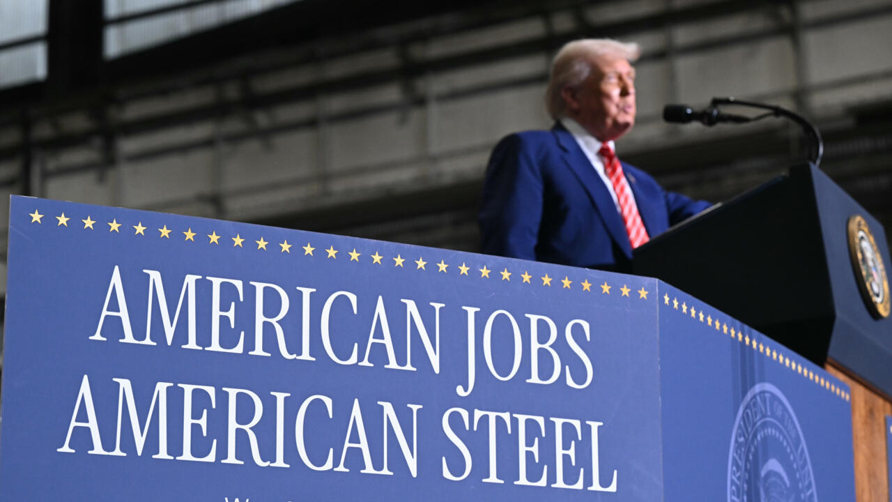 Person at a podium, beside a sign reading "American Jobs, American Steel," with a blurred industrial background.