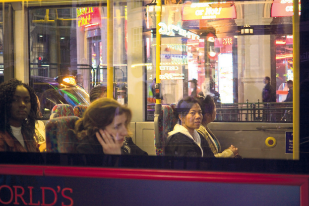 Passengers on a London double-decker bus at night, gazing out the window, with neon city lights reflecting on the glass.