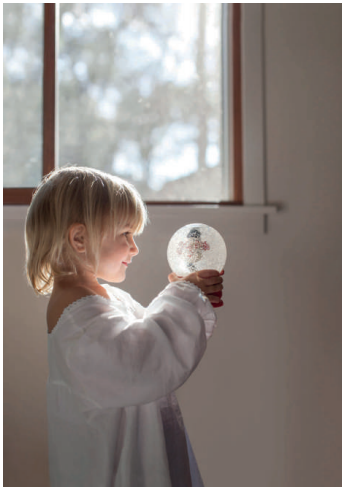Child in a white shirt gazing at a snow globe with a small snowman inside, standing by a window with soft daylight illuminating the scene.