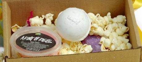 Cardboard box containing a tub of shower cream, a white bath bomb, a purple soap, and packing popcorn for padding.