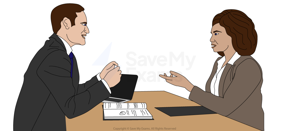 A man and woman in business attire are having a serious discussion at a table, with a laptop and open book in front of them.