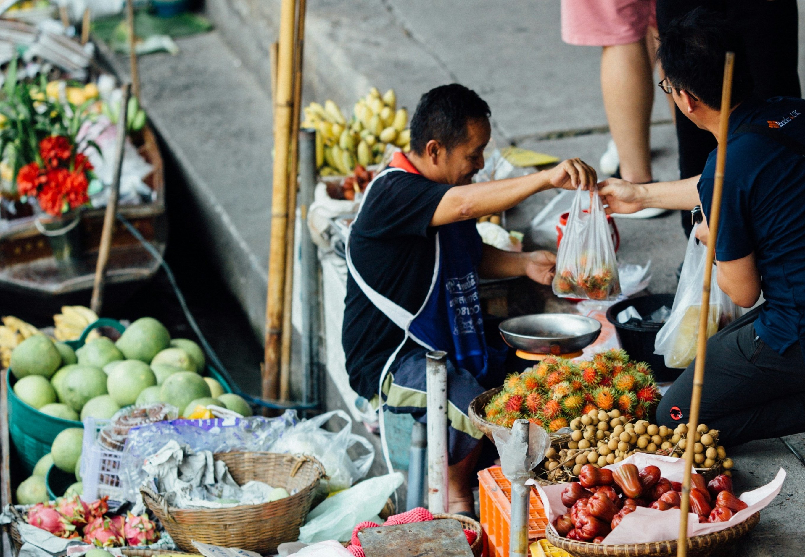 Market vendor handling rambutan in a plastic bag, surrounded by various tropical fruits, including mangoes and bananas, on a sidewalk.