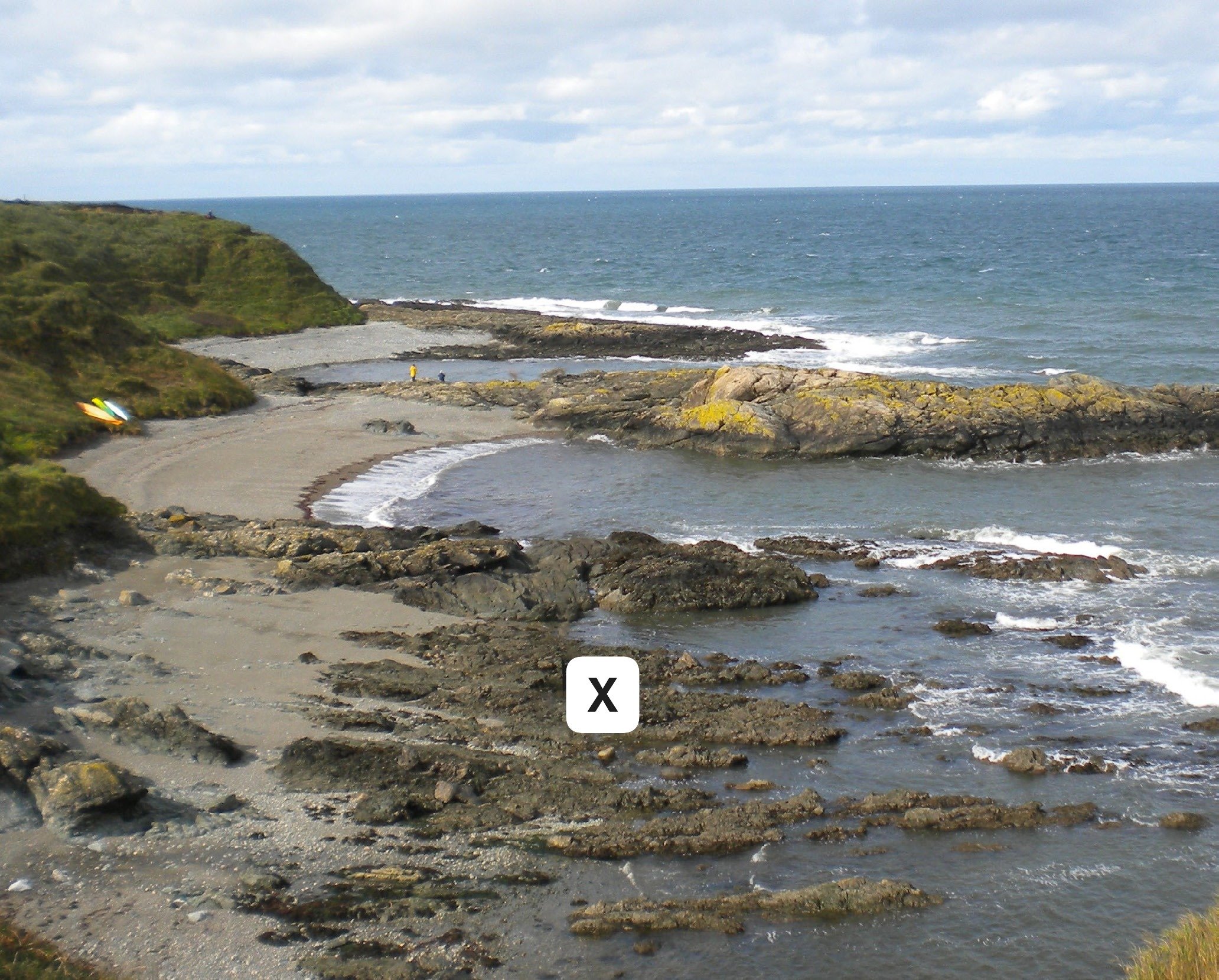 Rocky coastal bay with sandy beach and green cliffs, waves lapping against rocks, and small boats on the shore under a cloudy sky.