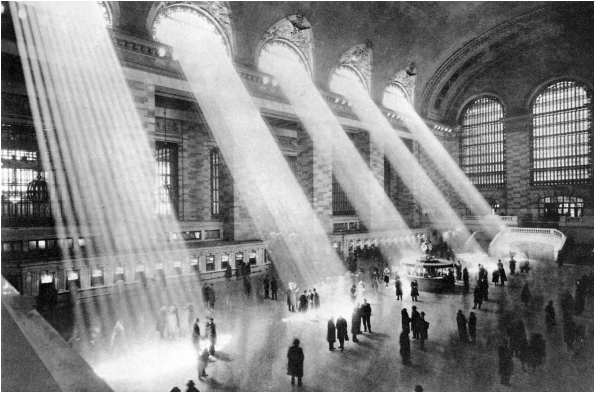 Sunlight streams through large arched windows, illuminating a busy train station with commuters walking on the marble floor and congregating around a kiosk.