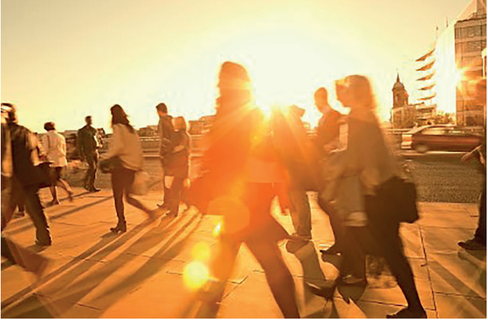 People walking briskly on a sunlit city street at sunset, with long shadows cast on the pavement and buildings partially visible in the background.