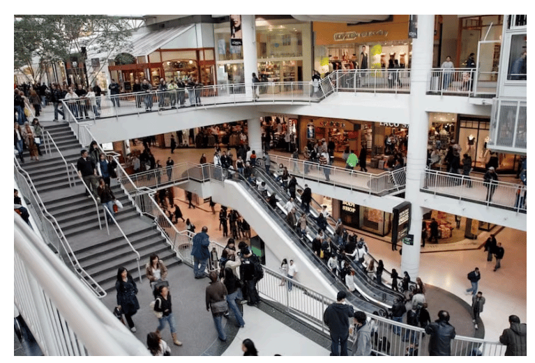 Busy shopping mall interior with multiple levels, escalators, and various shops. People are walking, using escalators, and standing around.
