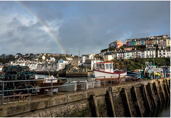 Harbour scene with boats docked, colourful houses on a hillside, a rainbow in a cloudy sky, and fishing nets on the quay.