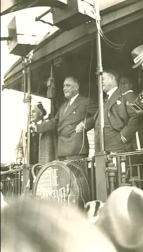 A man in a suit stands smiling on a train carriage platform, surrounded by others, with a round sign partially visible in front.