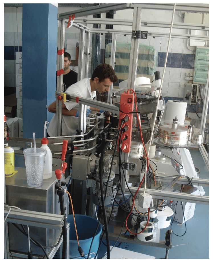 Man operating machinery with wires and bottles in an industrial setting, surrounded by tools, papers, and a blue bucket.