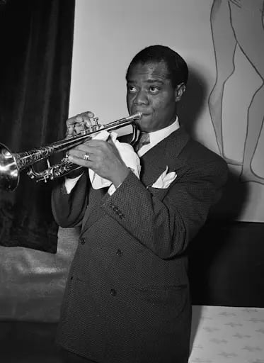 A man in a suit plays a trumpet indoors, with a curtain and artwork in the background, creating a classic jazz atmosphere.