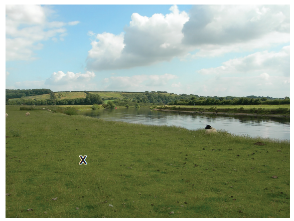 Rural landscape with a gentle river flowing alongside a grassy field under a partly cloudy sky, marked with an 'X' on the grass.
